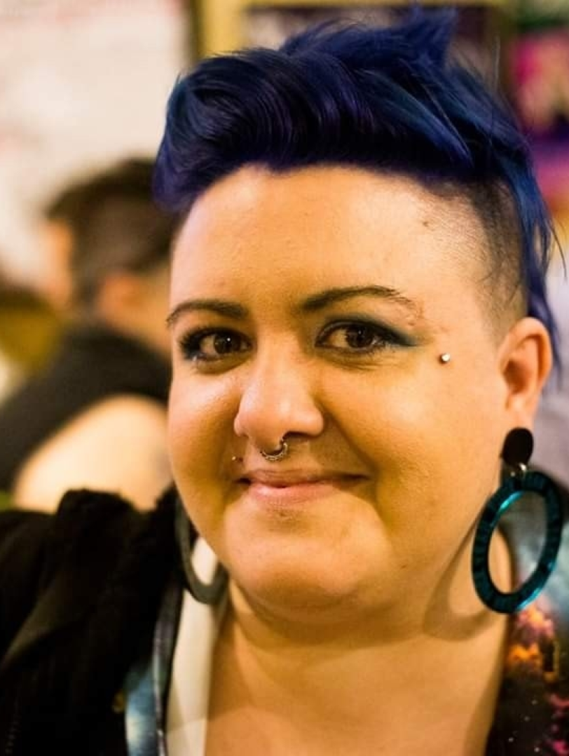 A picture of a non binary person with big round earrings and short blue hair smiling at the camera